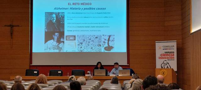 Una conferencia sobre el Alzheimer con dos ponentes y una presentación en una sala llena de público.