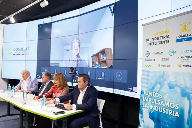 La imagen muestra un evento con varios ponentes en una mesa y una videoconferencia en una pantalla grande.