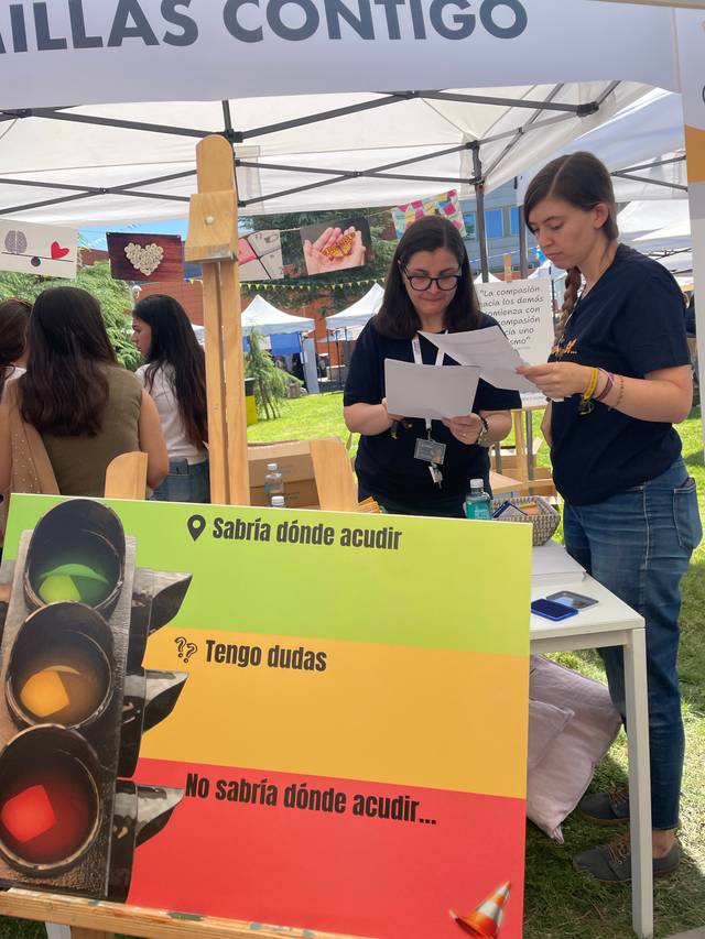 Dos mujeres revisan documentos en un stand con un cartel de semáforo que indica diferentes situaciones de incertidumbre.
