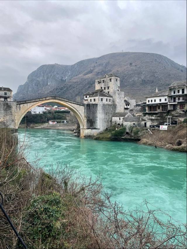 La imagen muestra un puente histórico sobre un río de color turquesa con montañas al fondo.
