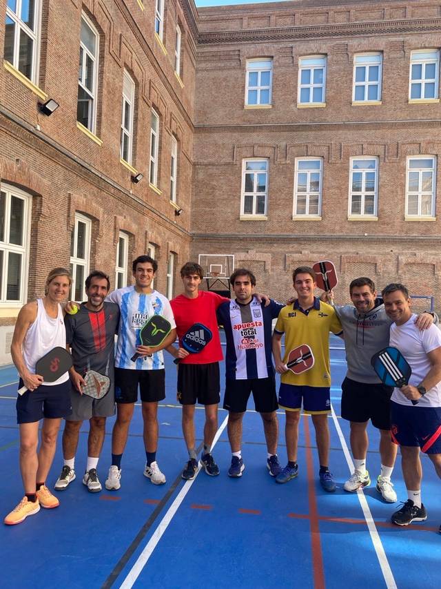 Un grupo de ocho personas sonrientes sosteniendo palas en una cancha de pádel al aire libre.