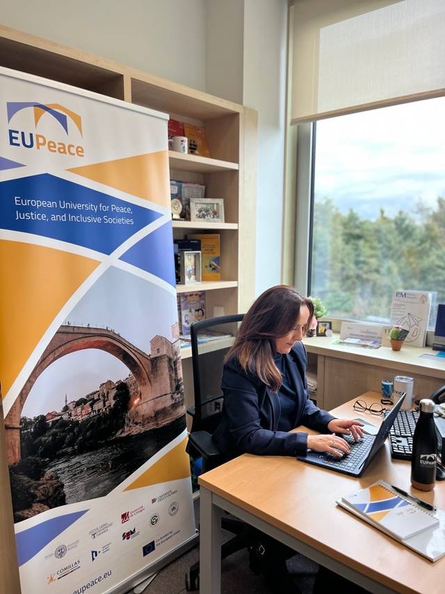 Una mujer trabaja en su escritorio frente a una ventana, con un banner de la Universidad Europea por la Paz detrás de ella.