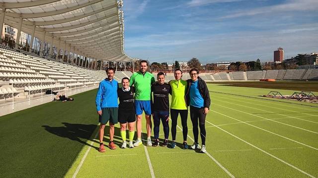 Un grupo de seis personas se encuentra en una pista de atletismo con un estadio al fondo.