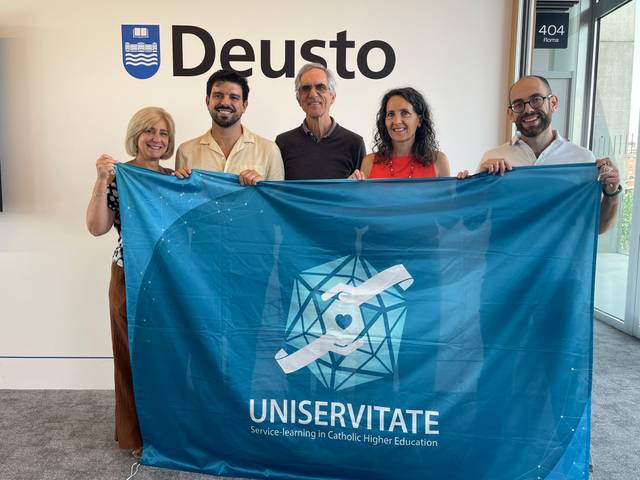 Un grupo de personas sosteniendo una bandera de UNISERVITATE en las instalaciones de la Universidad de Deusto.