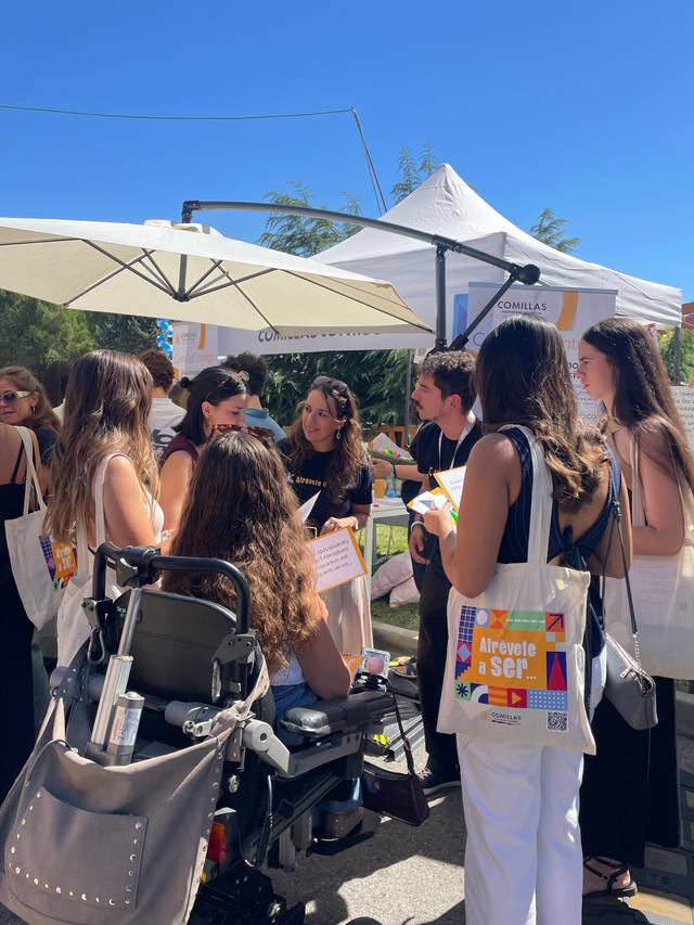 Un grupo de personas conversa en un evento al aire libre, rodeadas de carpas y con bolsas de tela.