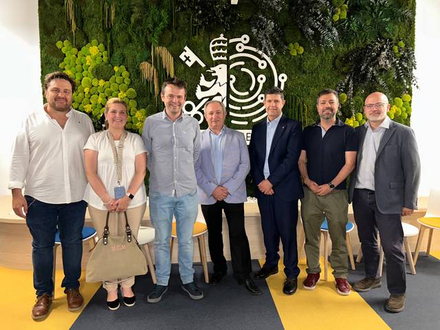 A group of seven people, consisting of six men and one woman, posing for a photo in a room with a green and white nature-themed backdrop and a logo.