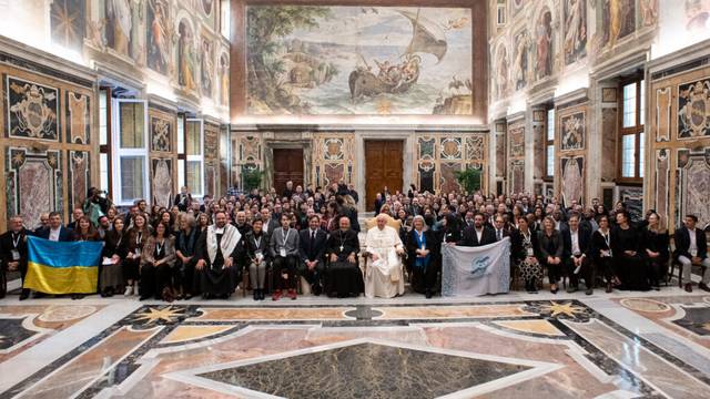 Un grupo numeroso de personas de pie en un salón adornado con frescos y mosaicos, posando para una fotografía con el Papa en el centro.