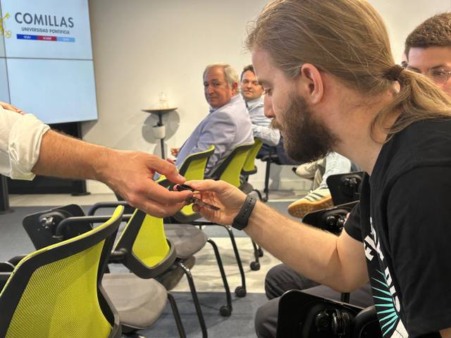 A man with long hair showing an electronic device to another person in a seminar room with people watching.