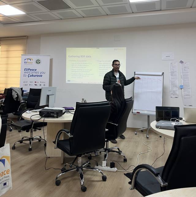 Una persona está presentando en una sala de trabajo con una pizarra y computadoras en el fondo.