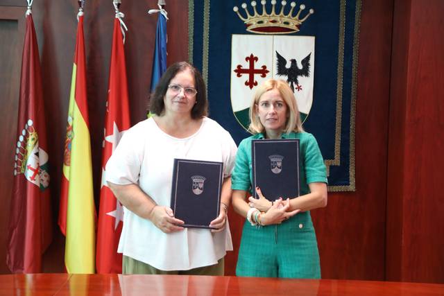 Dos mujeres sosteniendo documentos en un entorno formal con banderas de fondo.