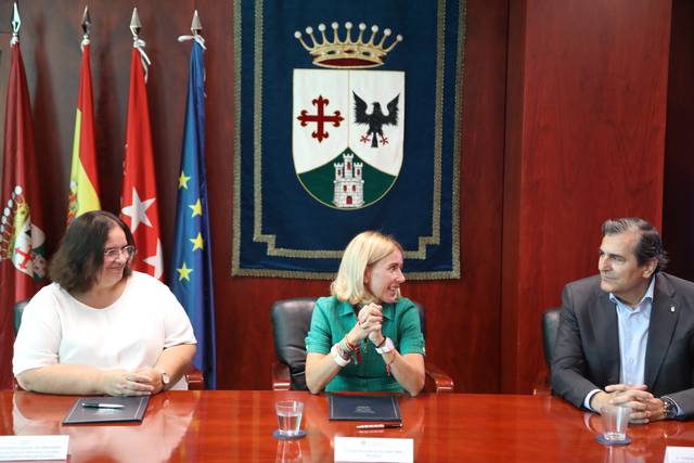 Una reunión formal con tres personas sentadas en una mesa frente a un fondo con banderas y un escudo.