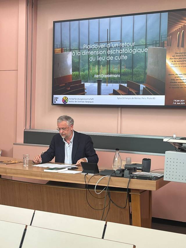 A man sitting at a desk in a lecture room with a presentation slide in the background.