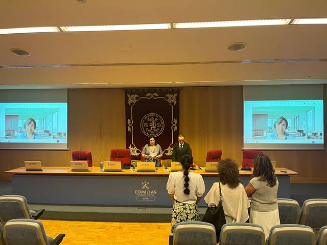 Una conferencia en una sala con presentadores y una pantalla que muestra a una persona.