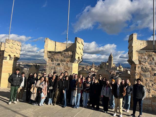 Grupo de personas posando para una foto en un puente histórico con vistas a una ciudad antigua bajo un cielo parcialmente nublado.