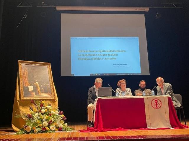 Una conferencia sobre espiritualidad femenina en el auditorio con un cuadro y decoraciones florales al fondo.
