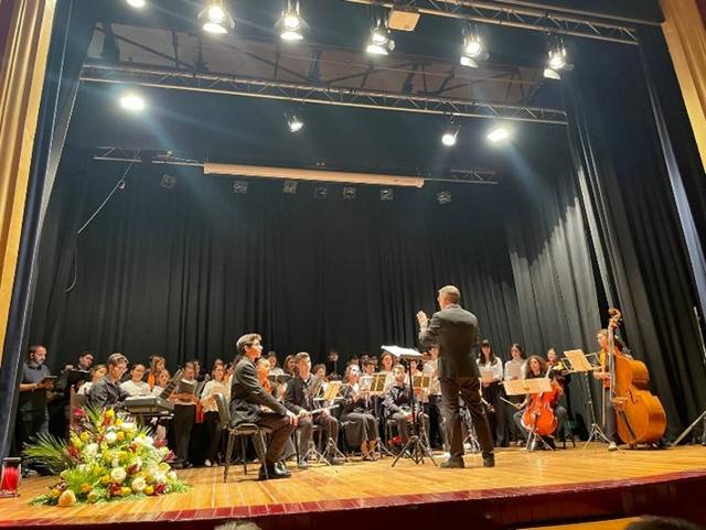 Una orquesta dirigida por un maestro en un escenario con músicos y un público visible.
