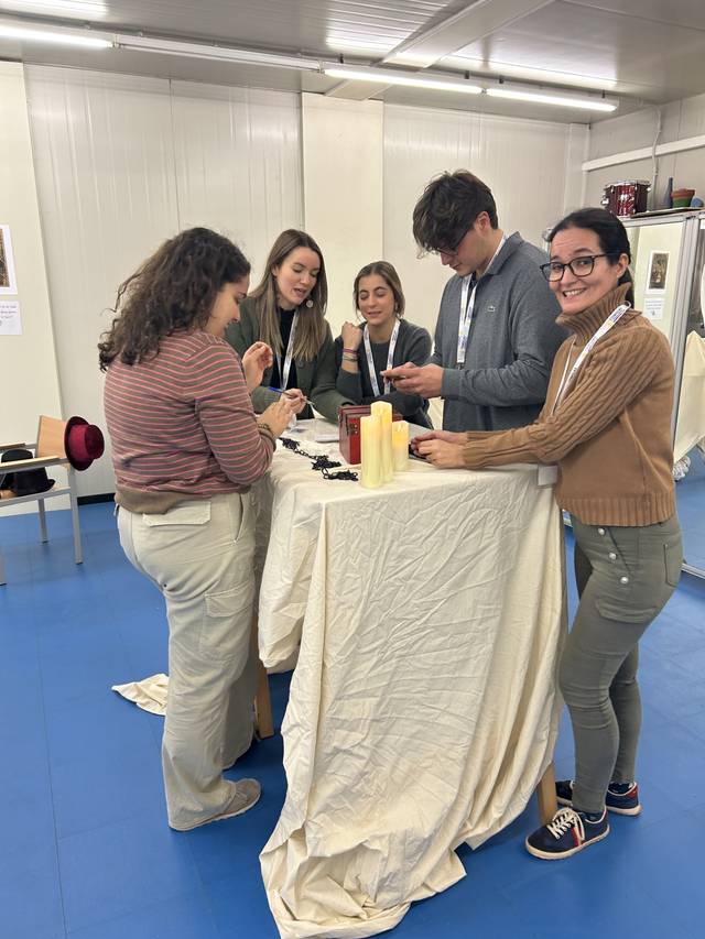 Un grupo de cinco personas jóvenes, incluyendo tres mujeres y dos hombres, están de pie alrededor de una mesa en una sala iluminada, participando en un taller o actividad de grupo.