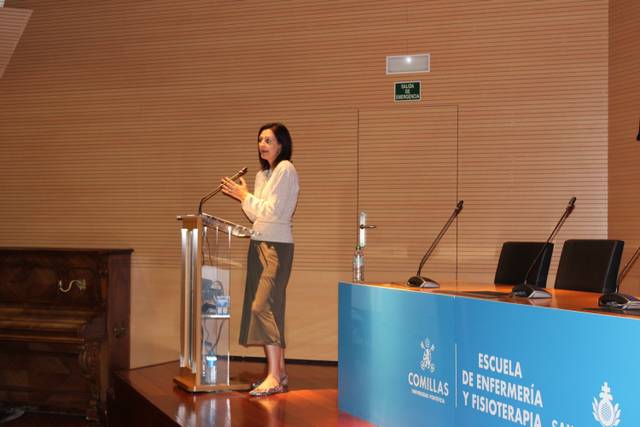 Una mujer está de pie detrás de un podio, hablando en un evento en una sala con un fondo azul que muestra el nombre de una escuela.