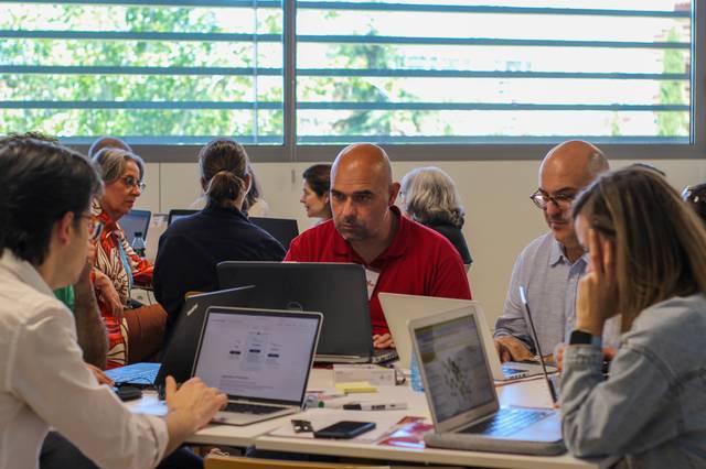 A group of adults engaged in a discussion at a table with laptops in a brightly lit room.