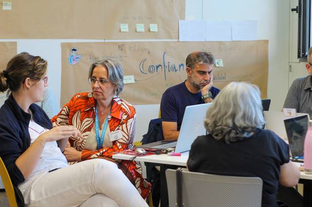 A group of adults sitting around a table, discussing and using laptops in what appears to be a workshop setting.