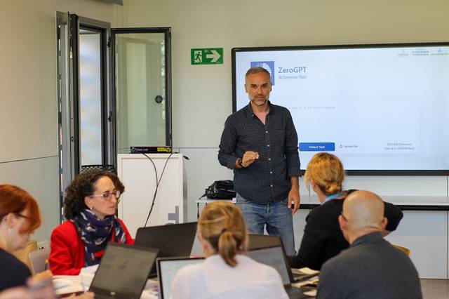 A man presenting in front of a digital screen to a group of seated adults in a classroom setting.