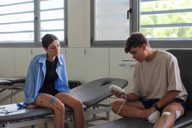 A young male athlete undergoing a muscle stimulation therapy on his leg, administered by a female physiotherapist in a clinical setting.