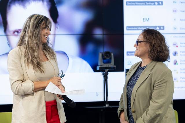 Dos mujeres conversan sonrientes en un evento, mientras una de ellas sostiene un micrófono y la otra lleva gafas.
