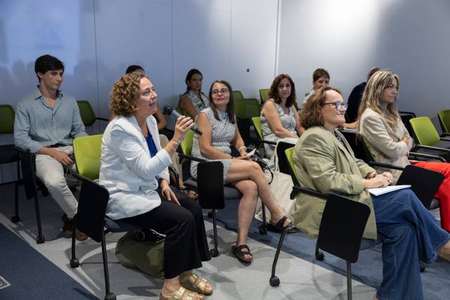 Un grupo de personas sentadas en un salón durante un evento o reunión, con una mujer levantando la mano para hacer una pregunta.
