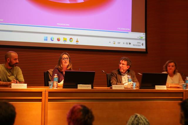 Four people sitting at a panel discussion in a conference room with a large screen displaying a colorful graphic in the background.