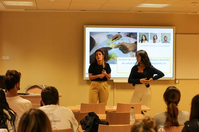 Two women are giving a presentation in a classroom with a slideshow in the background.