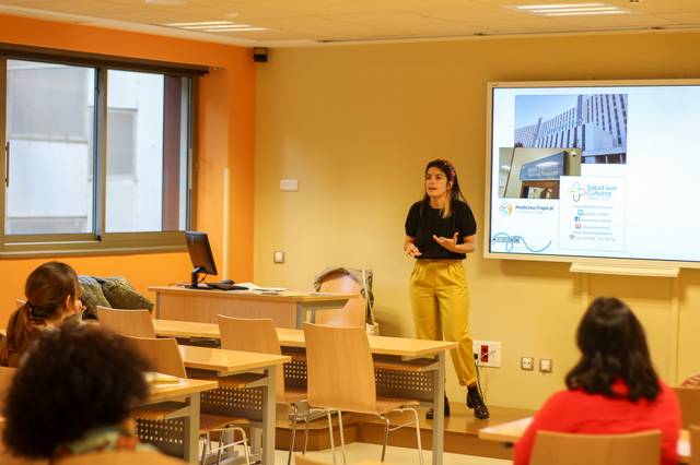 A woman presenting in a classroom with a digital screen showing a presentation behind her.