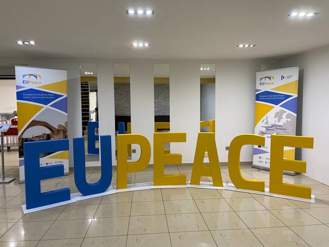 Una instalación de letras grandes que dicen 'EU PEACE' en un entorno interior con paneles informativos a los lados.