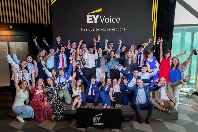 A group of people celebrating and posing for a photo in a room with a banner reading 'EY Voice, Haz que tu voz se escuche.'