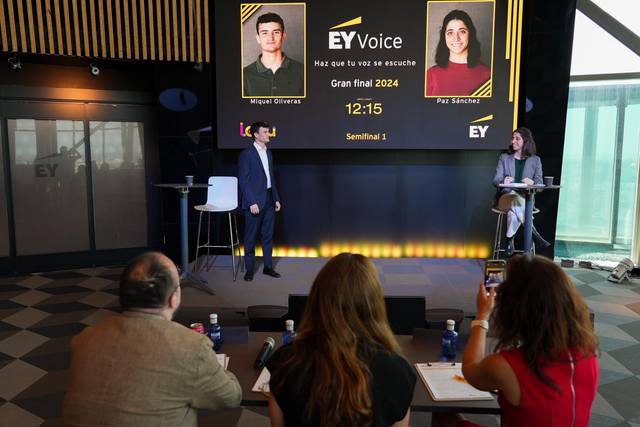 A man stands presenting on a stage with a large digital display behind him, showing profiles and the schedule of a speaking competition, while an audience watches.