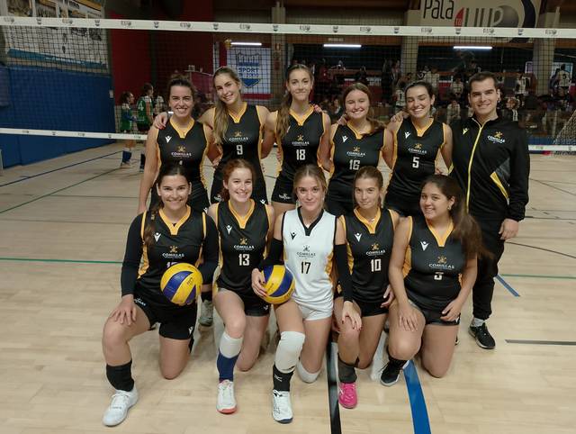 Un equipo de voleibol femenino posa sonriente en una cancha con uniformes negros y dorados.