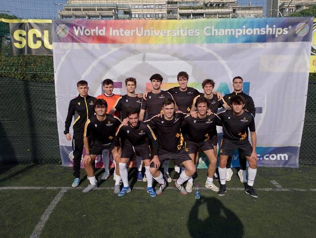 Un equipo de fútbol masculino posa en una cancha con un cartel de 'World InterUniversities Championships' al fondo.