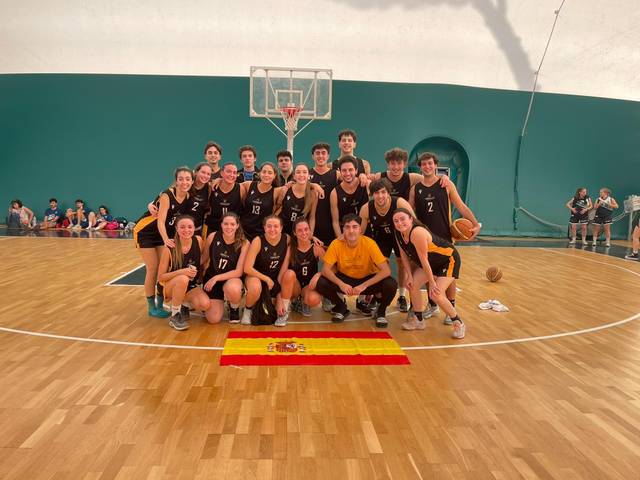 Un grupo de jóvenes atletas posan sonrientes en una cancha de baloncesto con una bandera en el suelo.