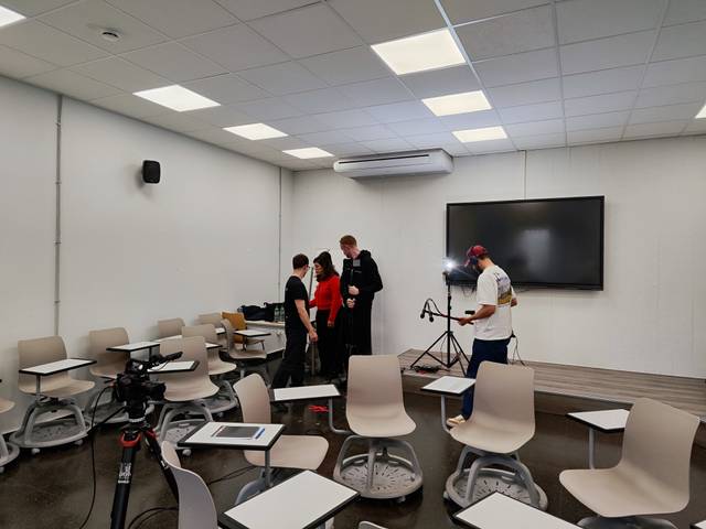 Un grupo de personas se prepara en un aula con escritorios y una pantalla grande al fondo.