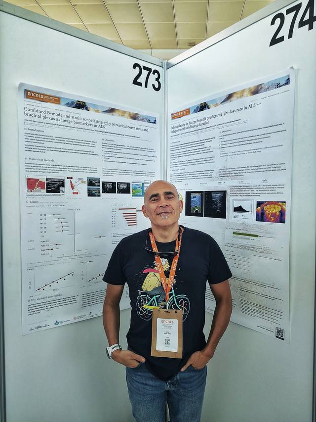 Un hombre sonriente se encuentra frente a dos paneles de presentación en un congreso científico.
