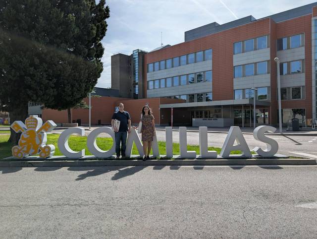 Dos personas posan frente al letrero de Comillas en un día soleado.