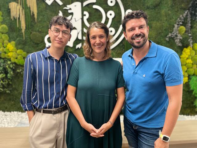 Tres personas posan juntas sonrientes frente a un fondo verde decorativo.