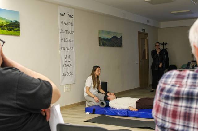 Una mujer está realizando una demostración de técnicas de relajación a otra persona en una sala, mientras un grupo de observadores asiste en el fondo.