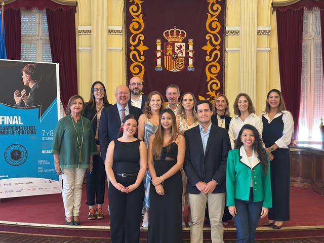 Un grupo de personas posando en un evento formal en un salón decorado con drapes y un escudo español.