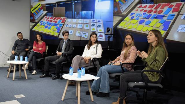 Un grupo de personas sentadas en una mesa durante un panel de discusión, con material visual colorido en la pantalla detrás.
