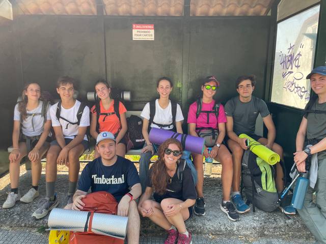 Un grupo de jóvenes con mochilas y esterillas esperan en una parada de autobús.