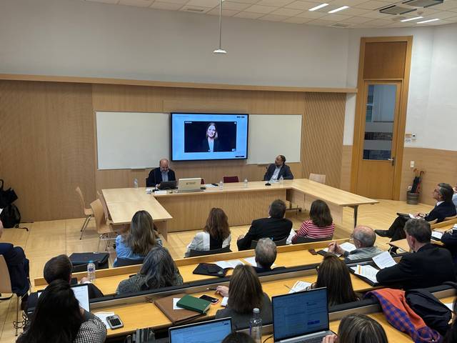 Un grupo de personas asiste a una conferencia en una sala, con una pantalla mostrando a un ponente virtual.