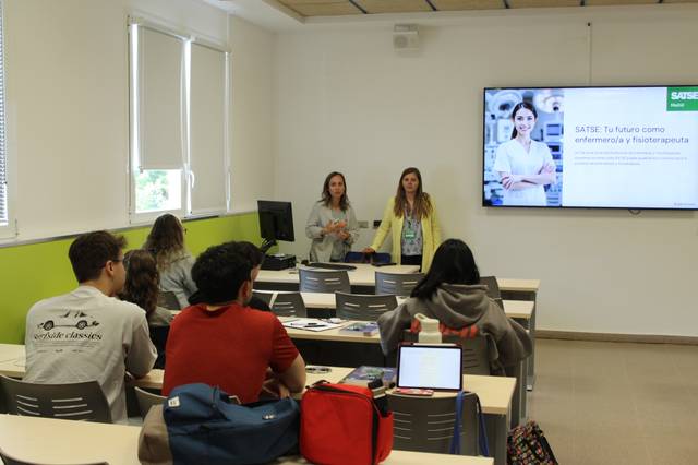Dos mujeres presentan un tema relacionado con la enfermería y la fisioterapia frente a un grupo de estudiantes en un aula.