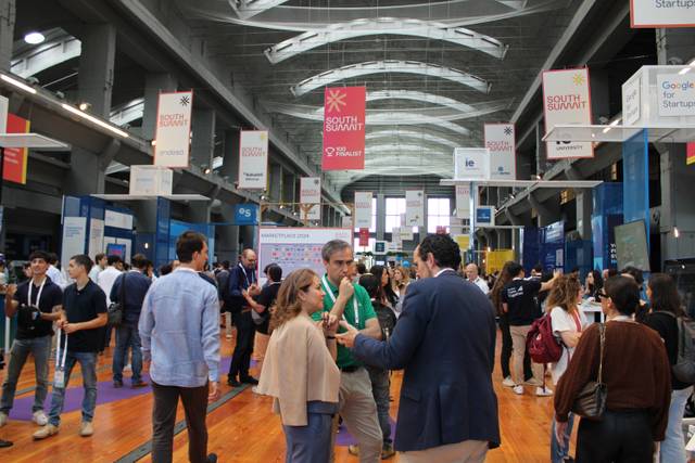 A bustling tech event with people engaging in discussions and networking in a large hall decorated with colorful banners.