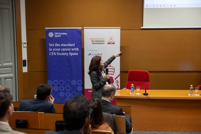 Una mujer presenta en una conferencia ante una audiencia sentada, señalando una pantalla en una sala de conferencias.