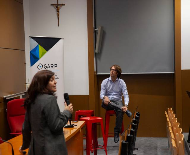 Una mujer está hablando con un micrófono en una sala de conferencias mientras un hombre se sienta y la escucha.